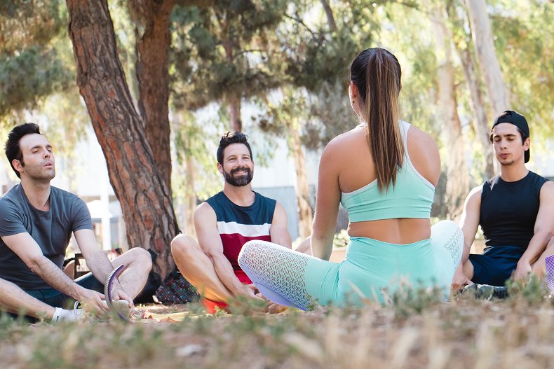 A yoga instructor in mint green attire leads a class with four male students sitting on the ground beneath tall trees in a park.